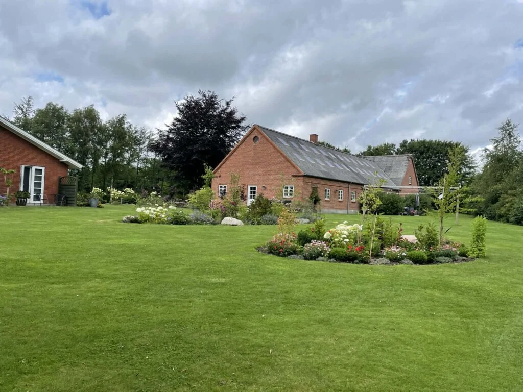 Huset set fra plænen: rødstenslænge med sort tag, bede og græs i forgrunden.